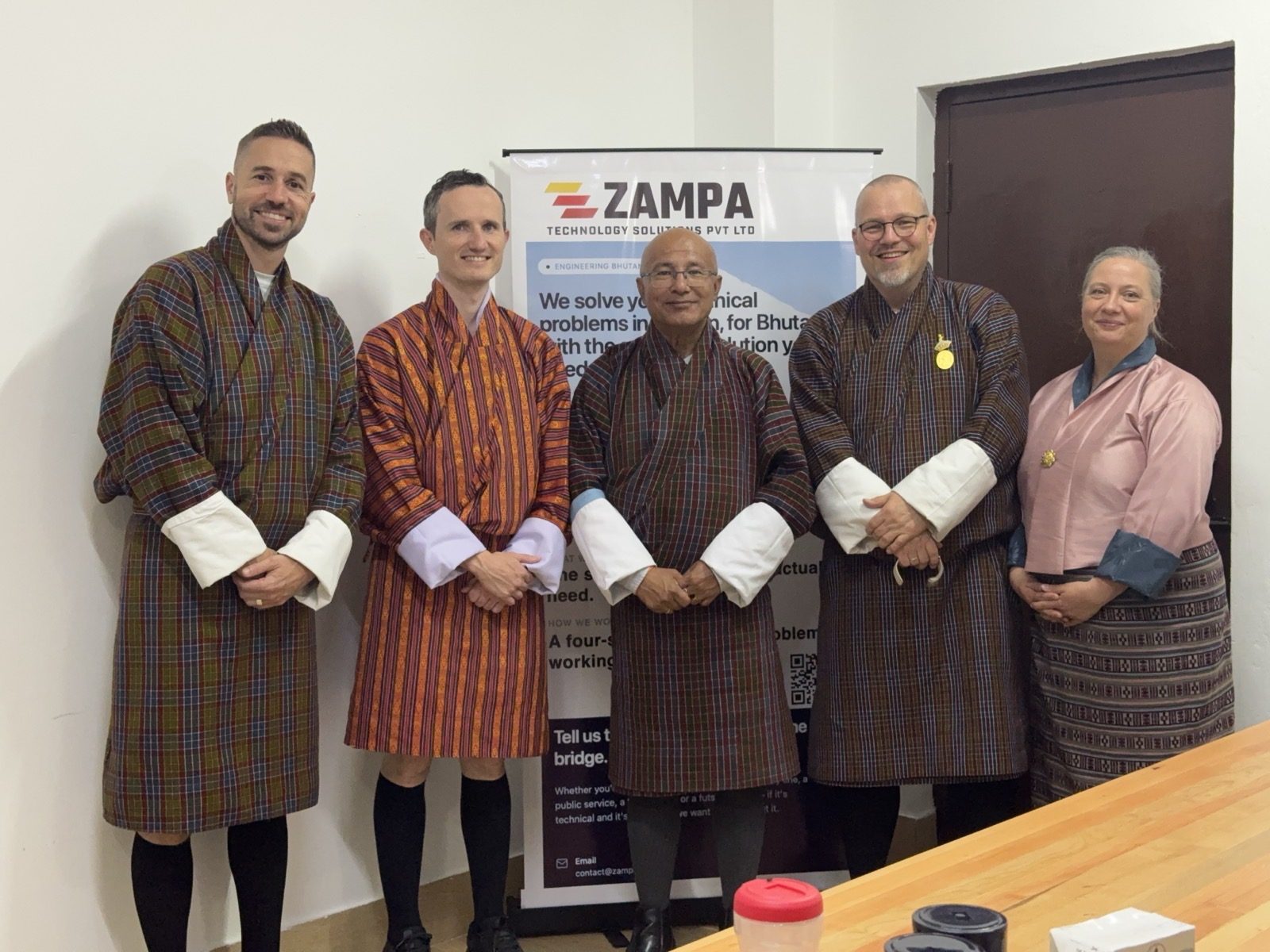 The Zampa team in gho and kira, standing with a Bhutanese colleague in front of the Zampa banner